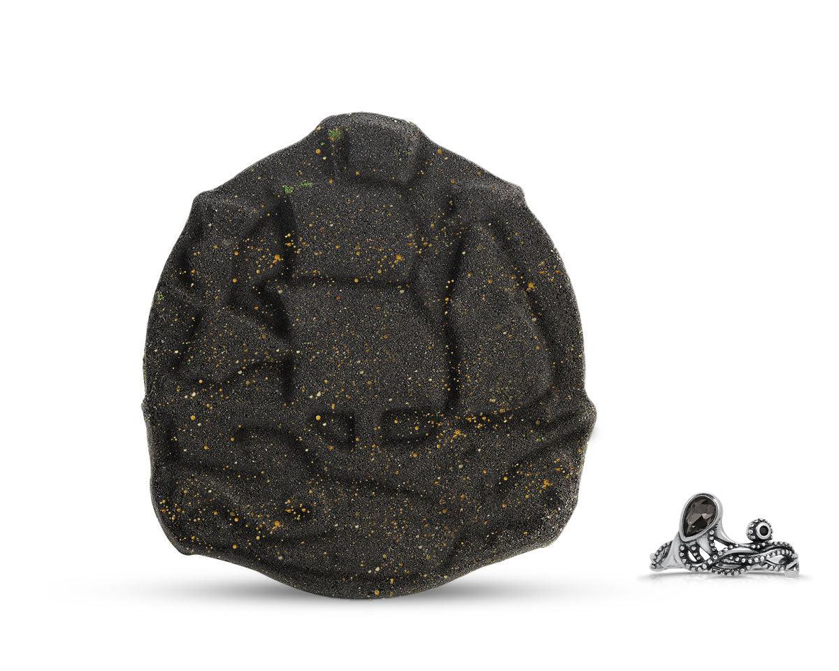 A black bath bomb with gold glitter, designed to resemble a pirate ship, with a small silver pirate ring beside it.